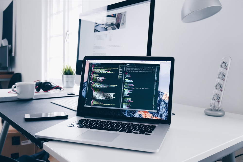 The-Basics A laptop displaying code sits on a desk next to a smartphone, headphones, and a monitor showing a webpage in a bright office setting.