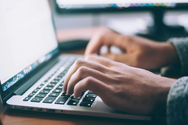 A person types on a laptop keyboard, with part of a computer monitor visible in the background.