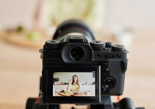 A digital camera records a woman sitting at a table with food, seen through the camera's viewfinder display, while filming a cooking scene.