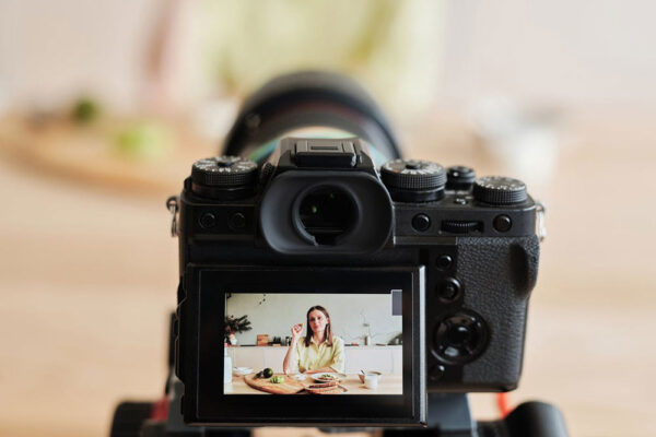 A digital camera records a woman sitting at a table with food, seen through the camera's viewfinder display, while filming a cooking scene.